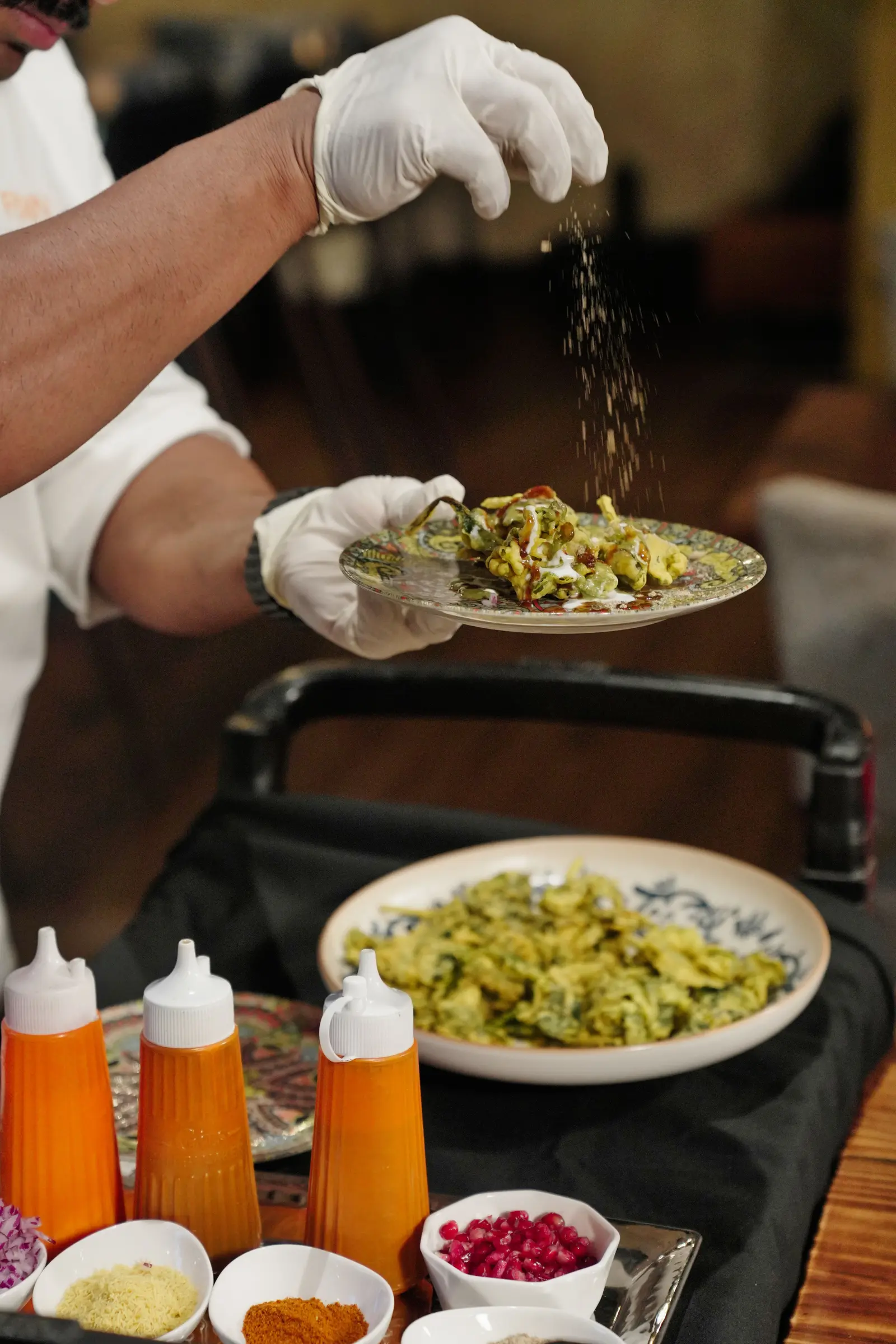 A chef's gloved hand holding a chaat plate as spice is sprinkled over it