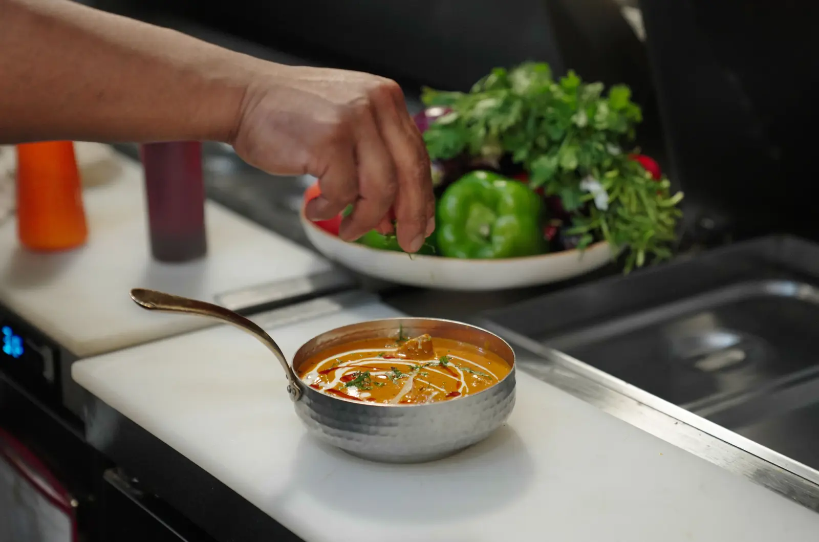 A curry in a hammered pan being finished at the pass, a bowl of herbs and peppers behind