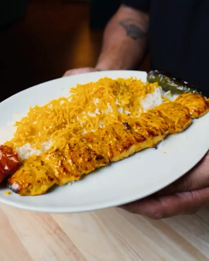 A chicken kebab on saffron rice with a grilled tomato and pepper, held out on a white plate